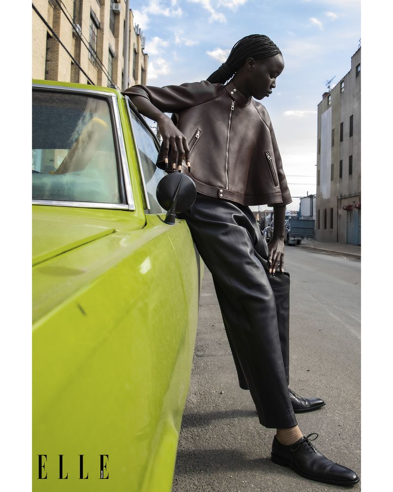 Model leaning against a bright green vintage car wearing a brown leather jacket and black trousers on a city street