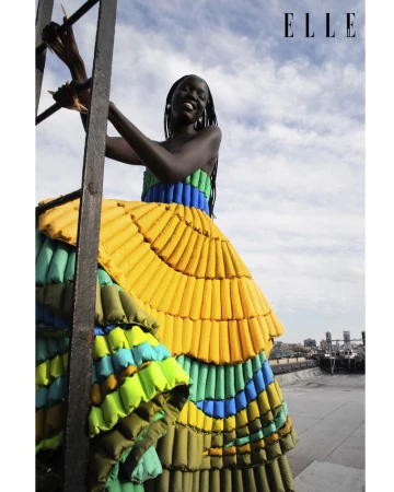 Model wearing a vibrant tiered puffer dress climbing a metal rooftop ladder against a cloudy sky
