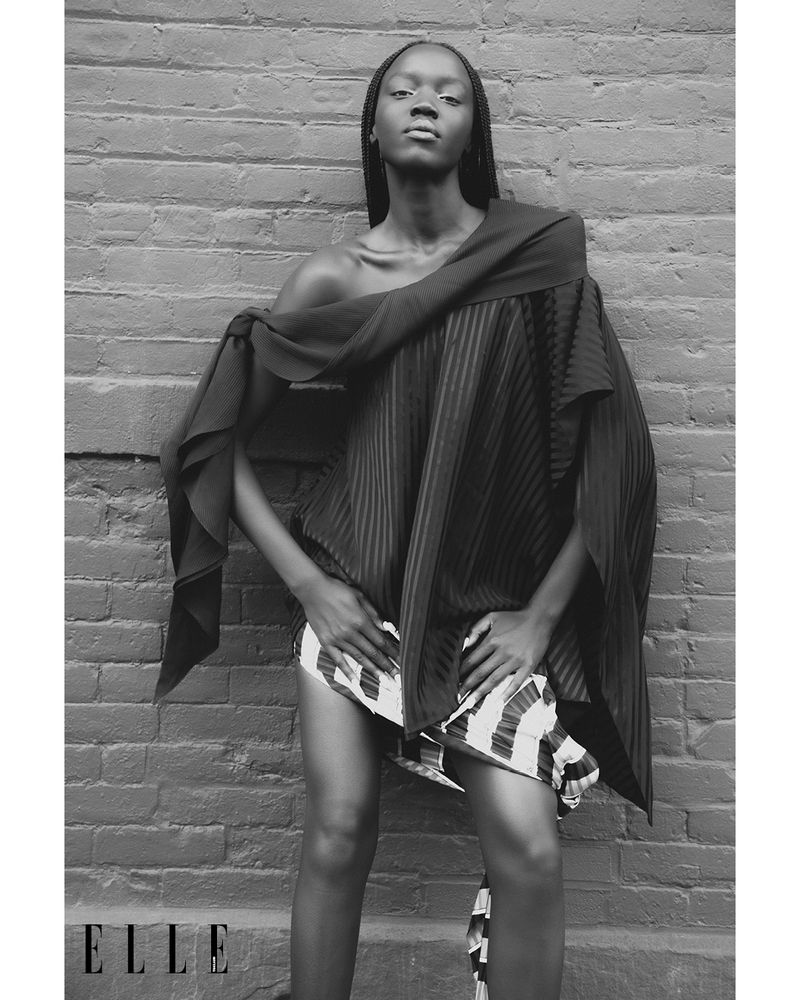 Black and white fashion shot of a Black female model posing against a brick wall, wearing a draped outfit. Elle logo visible.