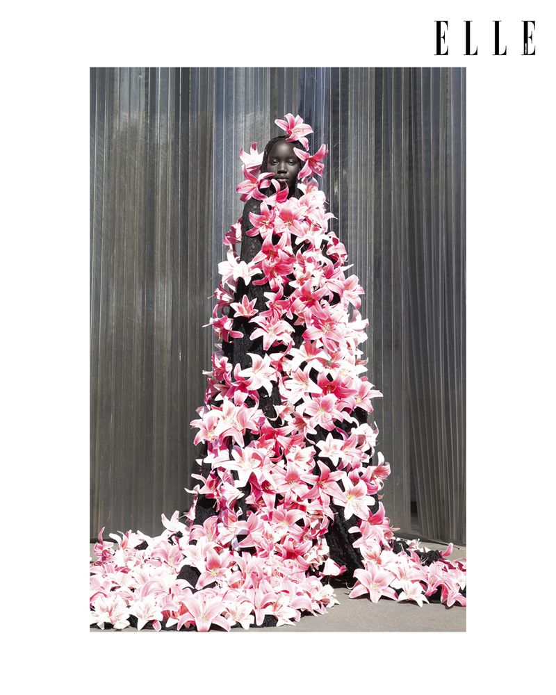 Fashion shot of a Black woman adorned with pink lily flowers, posing against a textured gray backdrop, as featured in Elle magazine.