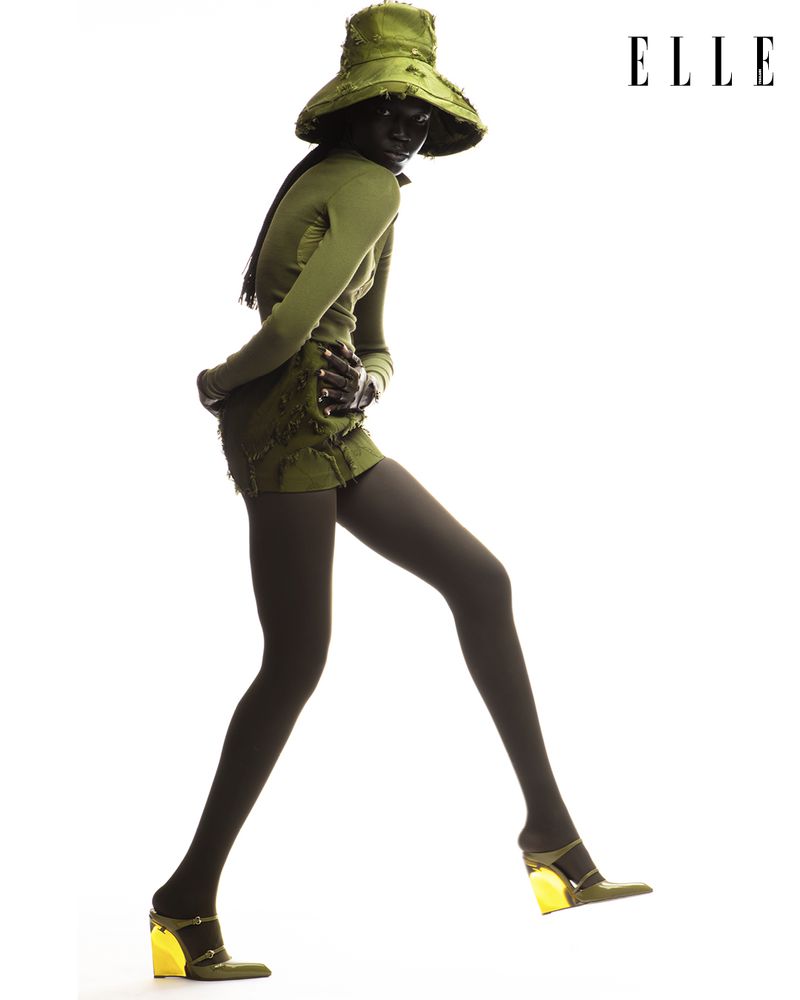 Fashion editorial shot of a Black female model wearing an olive green ensemble with a floppy hat and gold-heeled shoes against a white background.