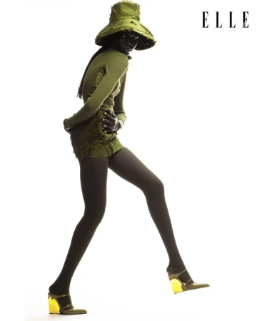 Fashion editorial shot of a Black female model wearing an olive green ensemble with a floppy hat and gold-heeled shoes against a white background.