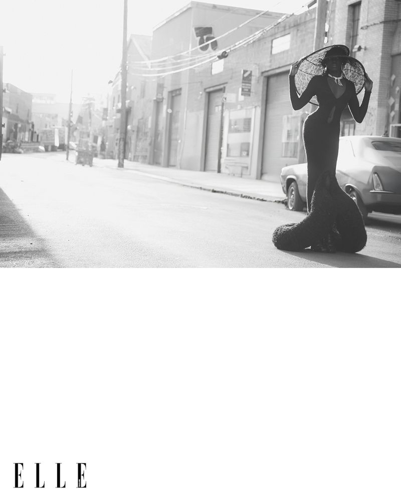 Elegant woman in black gown and wide-brimmed hat poses on city street with classic car and industrial buildings in a black and white editorial.