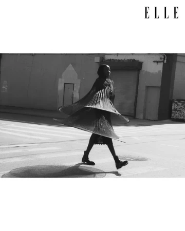 Black and white fashion editorial shot of a Black female adult walking across a crosswalk in a dress with a pleated circular skirt.
