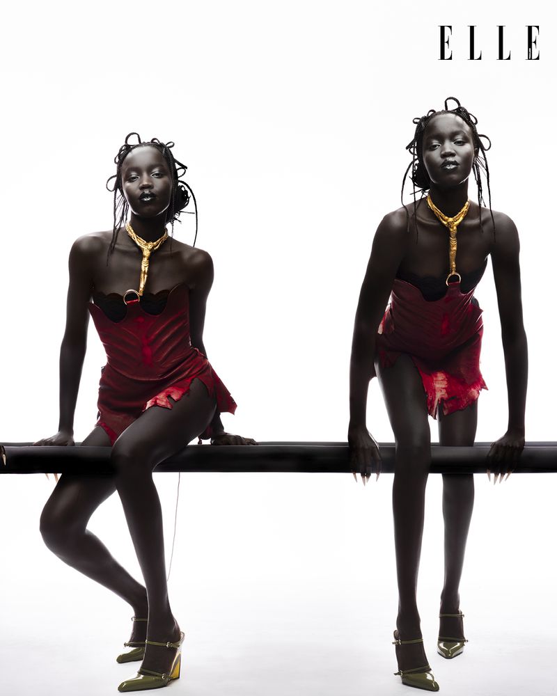 Two Black female models in red dresses and gold jewelry pose in a studio against a white backdrop. The women wear gold high heels.