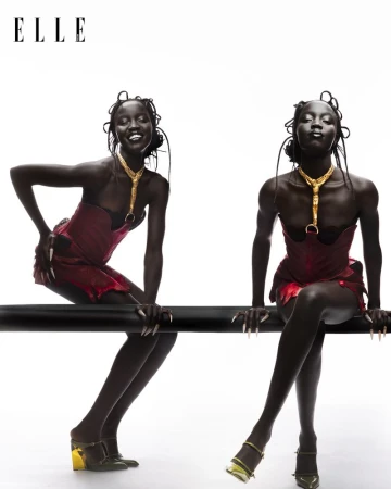 Two women with dark skin and elegant gold jewelry posing in red corset dresses and heeled shoes, sitting on a black bar on white.