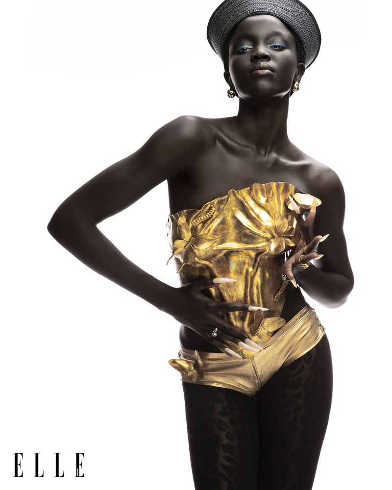 A dark-skinned woman poses confidently in gold petal-like swimwear and a dark hat against a bright white background in a fashion shot.