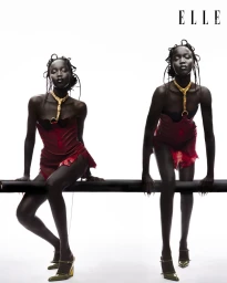 Two black female models wearing red corsets and gold necklaces seated on a dark rail against a bright white background for Elle Thailand.