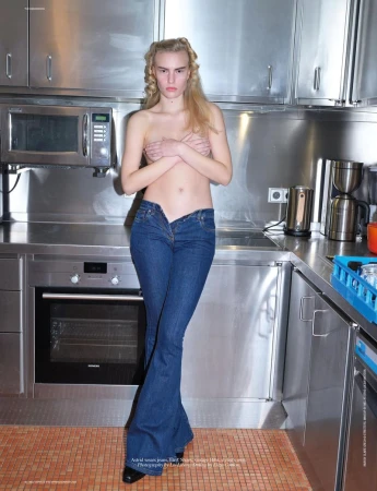 A female model stands in a kitchen wearing jeans with an open fly, her chest covered by her arms in a fashion editorial style.