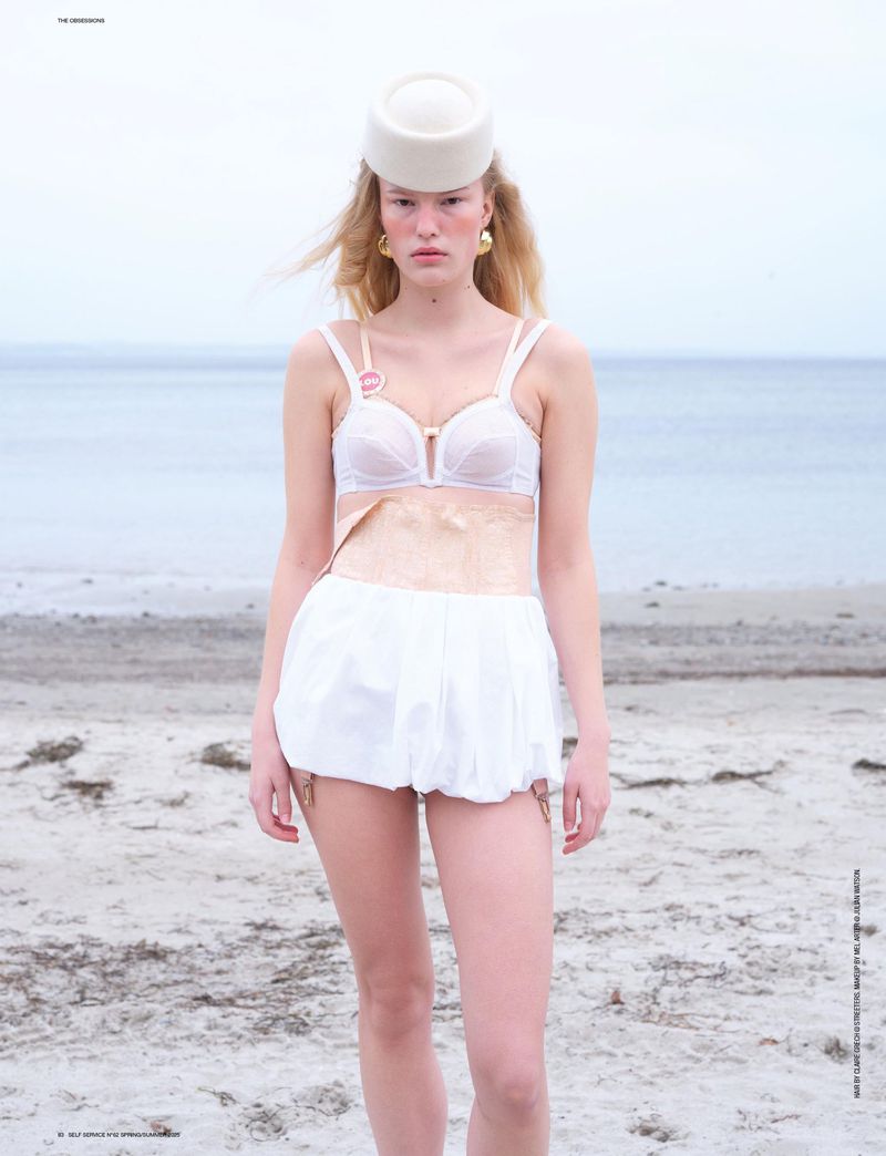 Blonde woman in lingerie and hat stands on beach with ocean background, showcasing fashion and editorial style in outdoor setting.