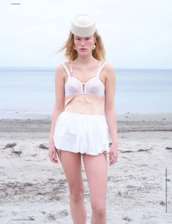 Blonde woman in lingerie and hat stands on beach with ocean background, showcasing fashion and editorial style in outdoor setting.
