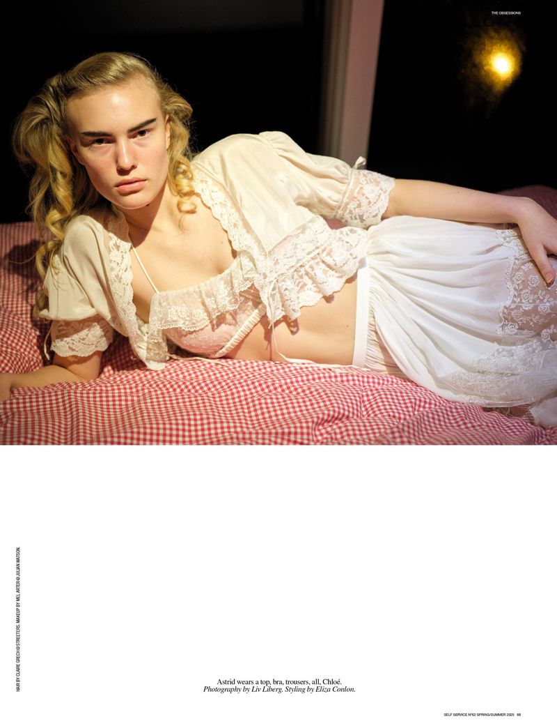 A fair-skinned woman with blonde hair lies on a bed with red gingham sheets wearing a white, lace-trimmed lingerie set and robe indoors.