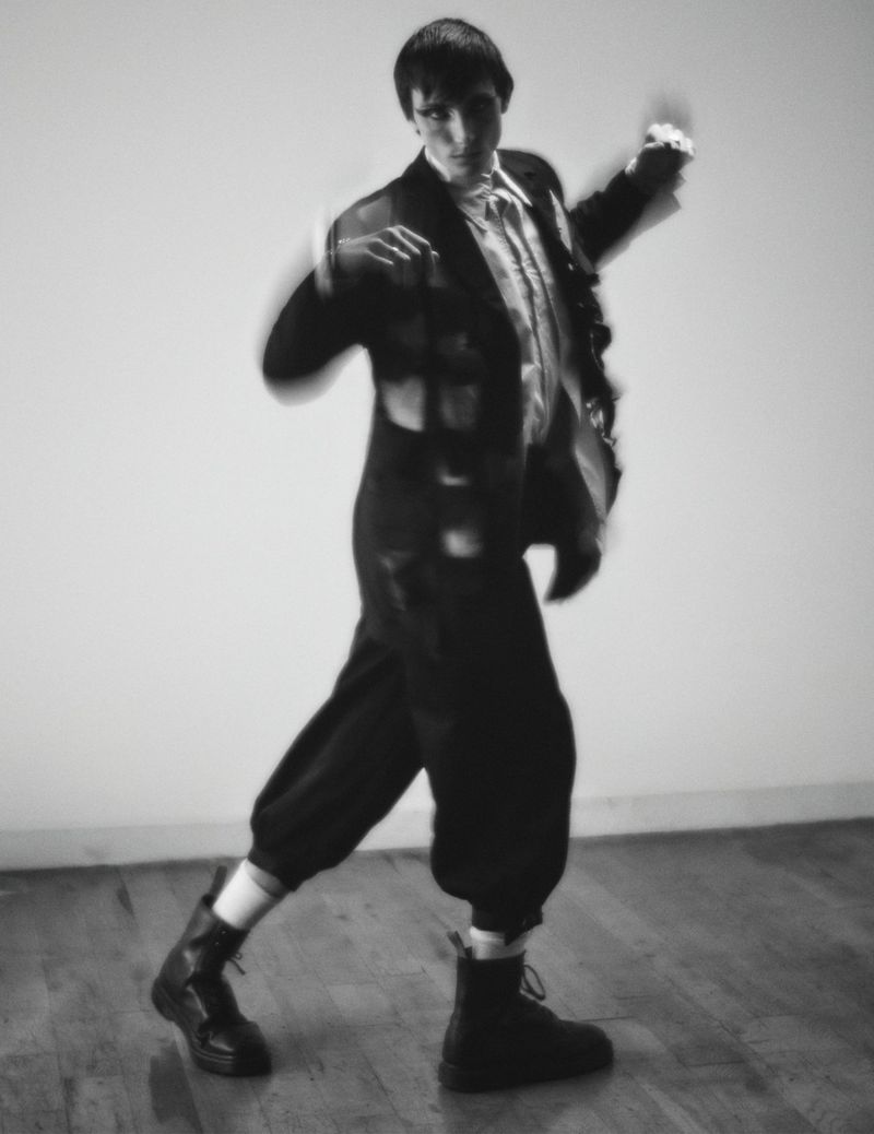 Full body shot of androgynous person in a black suit with boots indoors against a pale wall, captured with motion blur.