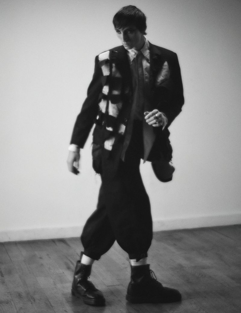 Monochrome portrait of a man in an ornate black suit jacket and black trousers, walking across a hardwood floor in a photo studio.