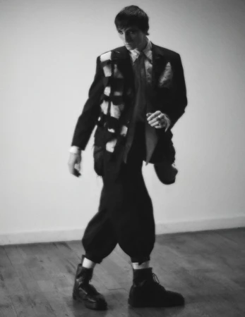 Monochrome portrait of a man in an ornate black suit jacket and black trousers, walking across a hardwood floor in a photo studio.