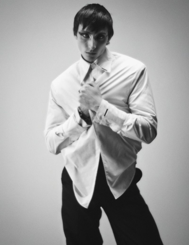 Monochrome portrait of a male model in a white button-down shirt and dark trousers against a plain background.