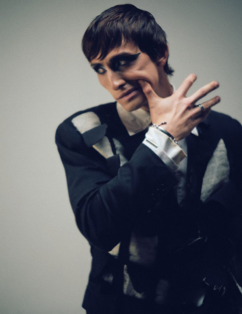 Man with dramatic dark eye makeup pushing his cheek inward with his fingers, wearing a dark jacket against a plain wall.