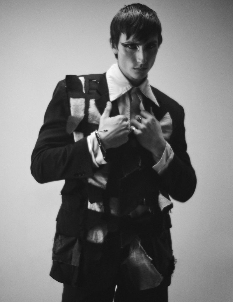 Black and white portrait of a slender adult man in a dark suit jacket, white collared shirt, with dark eye makeup, indoors.