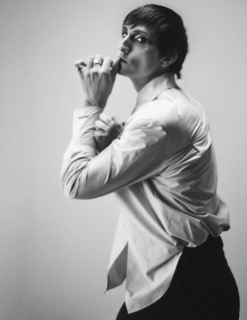 Portrait of a person in a white shirt with short dark hair, posed with hands near the face, in a black and white photo studio setting.