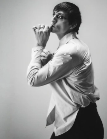 Portrait of a person in a white shirt with short dark hair, posed with hands near the face, in a black and white photo studio setting.