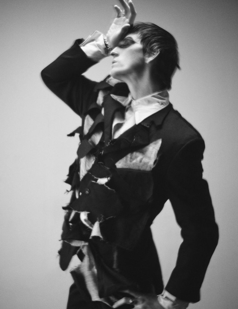 Studio shot of person in avant-garde fashion, wearing a textured black jacket and white collared shirt, posing against a grey backdrop.