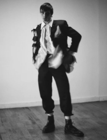 Model standing in dark trousers, white shirt, and jacket against a plain wall and wood floor in a black and white editorial photo.