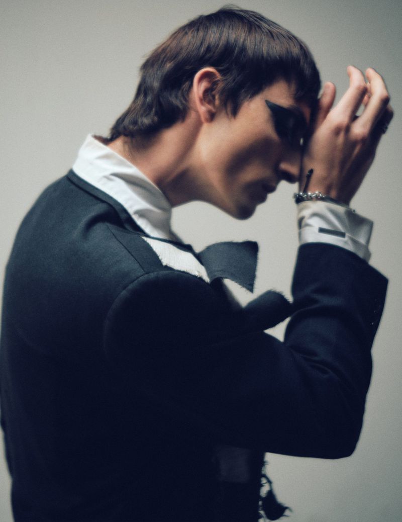 Stylish male model with dark eye makeup and retro hairstyle poses in a studio wearing a black and white suit jacket.