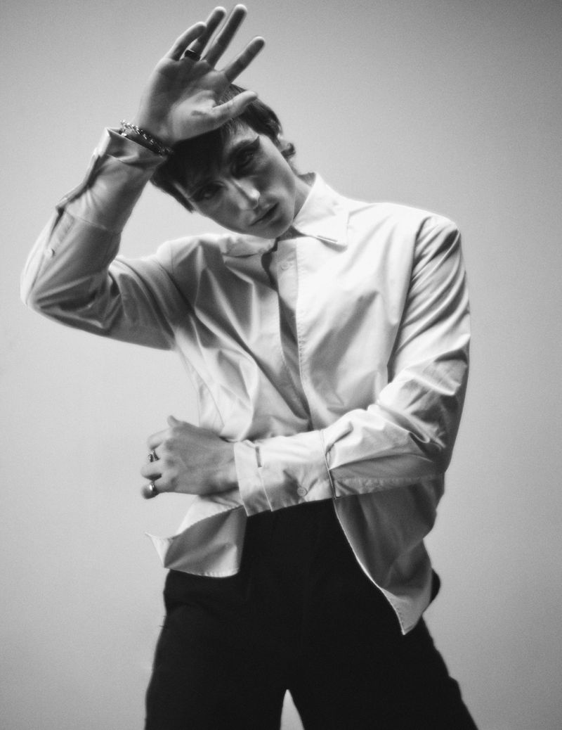 Black and white portrait of a person in a white button-down shirt posing with hand raised to forehead.