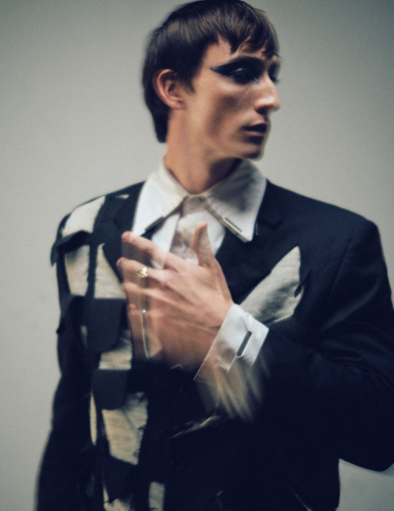 Stylish adult male model in dark blazer with geometric patches, white dress shirt, and heavy makeup, looking left in a studio shot.