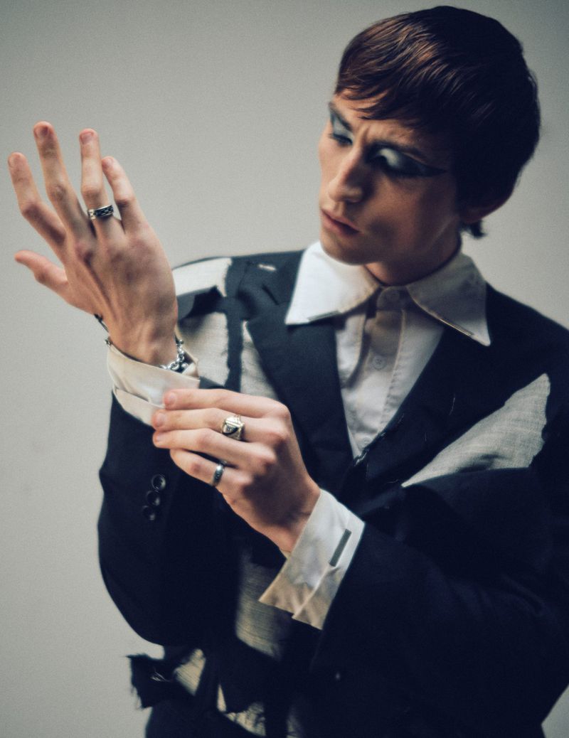 Man adjusting his suit cuff wearing silver rings and dramatic eye makeup against a white background