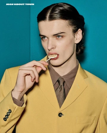 Close-up of a young man with dark hair and blue eyes, wearing a yellow suit and brown shirt, holding a lollipop against a teal background.