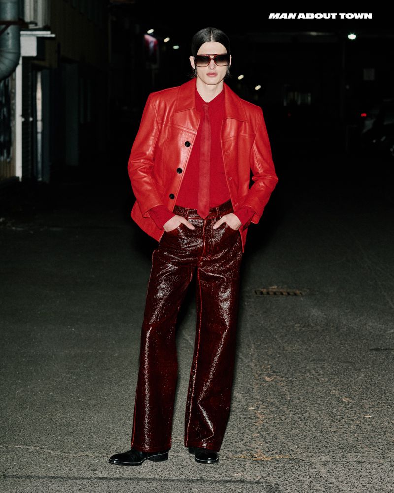 Fashionable man poses in red leather jacket and glossy brown pants on a city street at night, highlighted by the contrast of his stylish attire.