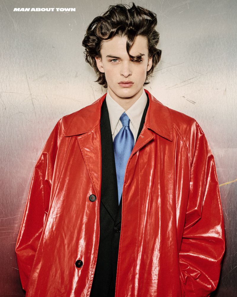 Man with curly dark hair wearing a red patent leather trench coat, black suit, and light blue tie posing against a silver background.