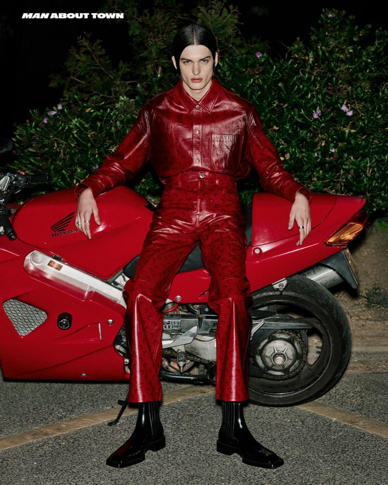 A male model in red leather-like outfit sits on a red Honda motorcycle at night in a fashion editorial photograph, with a dark background.