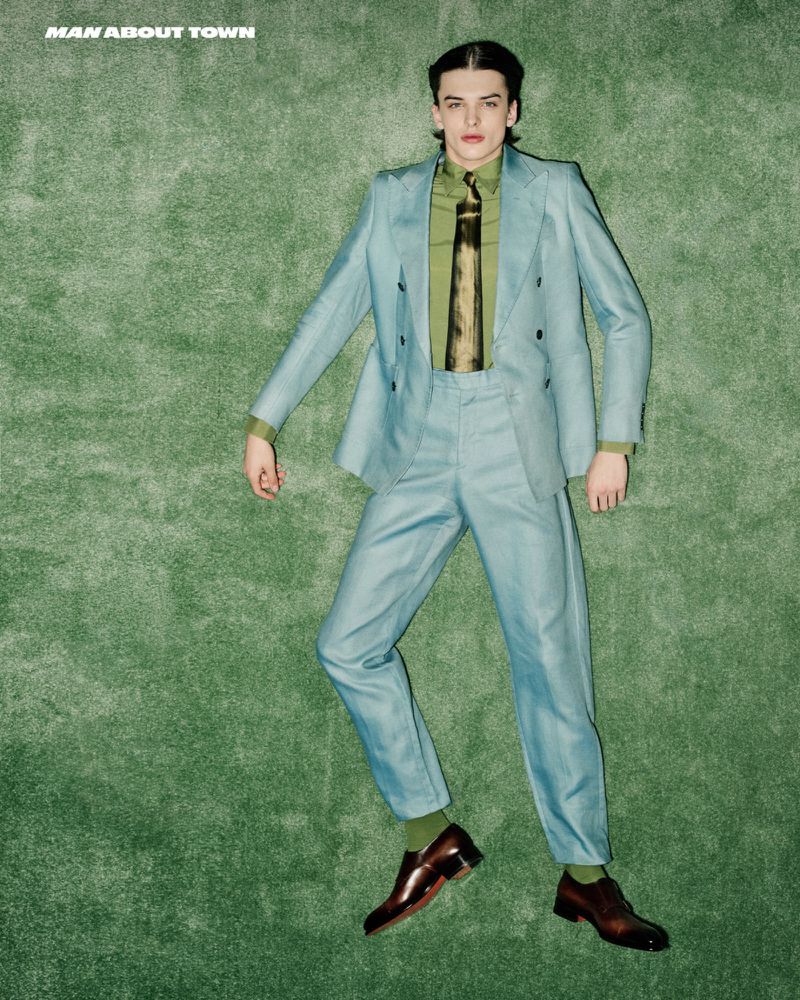 A young adult male poses in a light blue suit on a textured green background. He wears a green shirt and patterned tie with brown dress shoes.