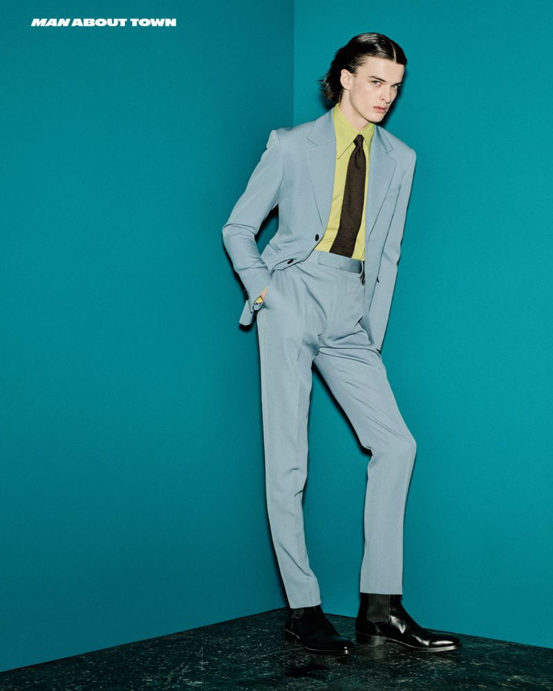 Man wearing a light blue suit and lime green shirt standing against a teal wall for Man About Town editorial
