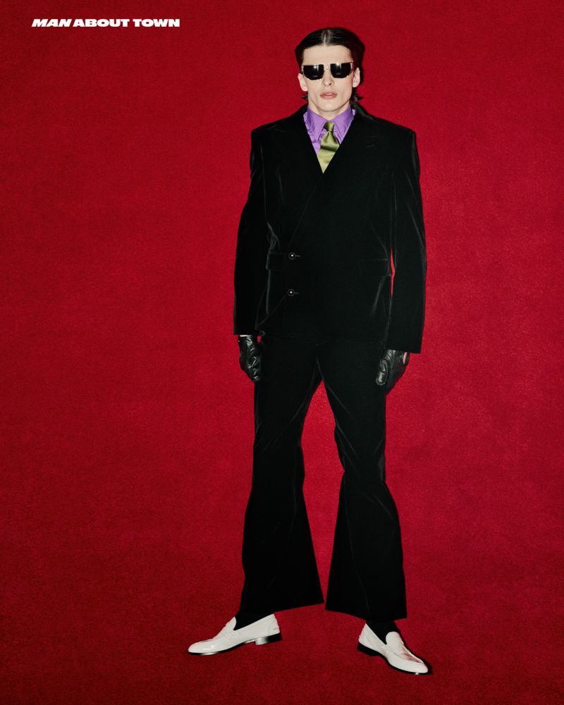 A man in a black velvet suit and sunglasses poses against a red background, showcasing retro-inspired fashion with white loafers and a lavender shirt.
