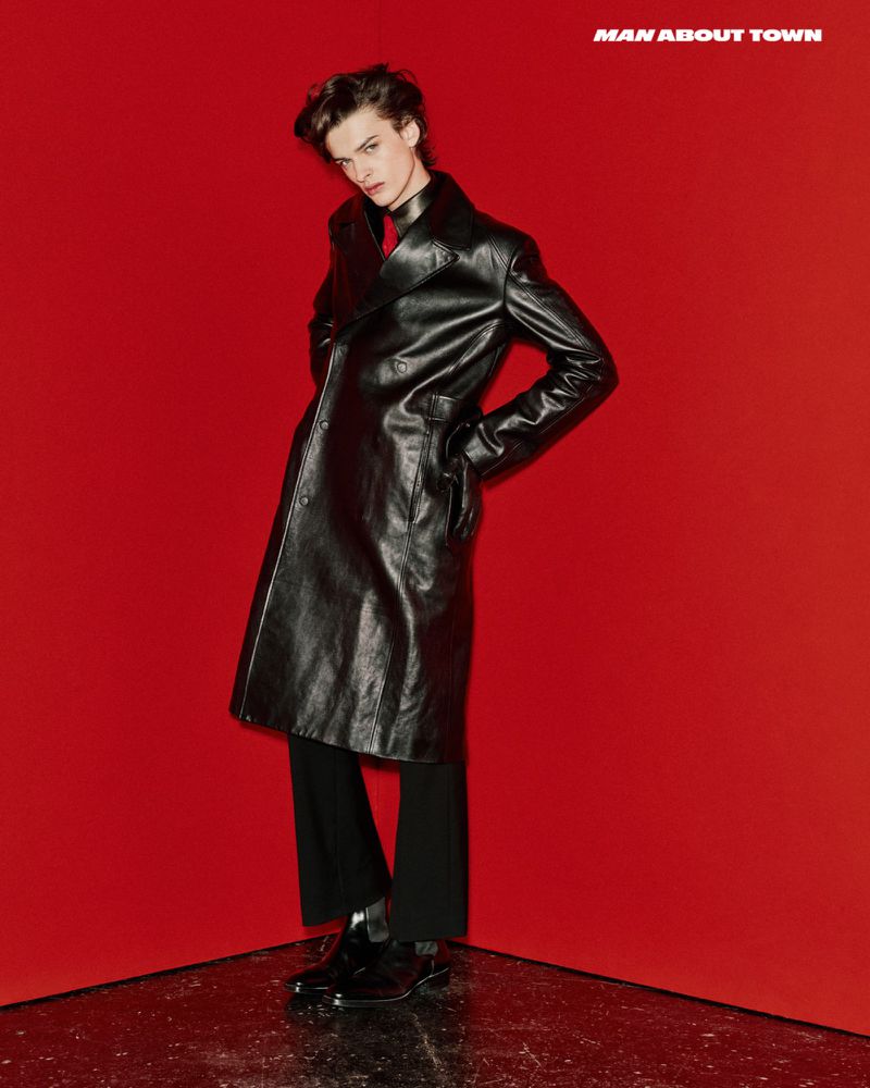 A young adult male model in a black leather coat poses against a red backdrop in a studio, showcasing a high-fashion editorial look.