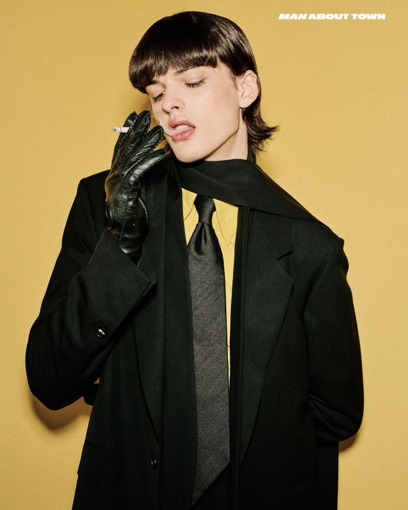 A young adult model in a black suit and gloves smokes a cigarette against a yellow background, editorial fashion shoot, Man About Town magazine.