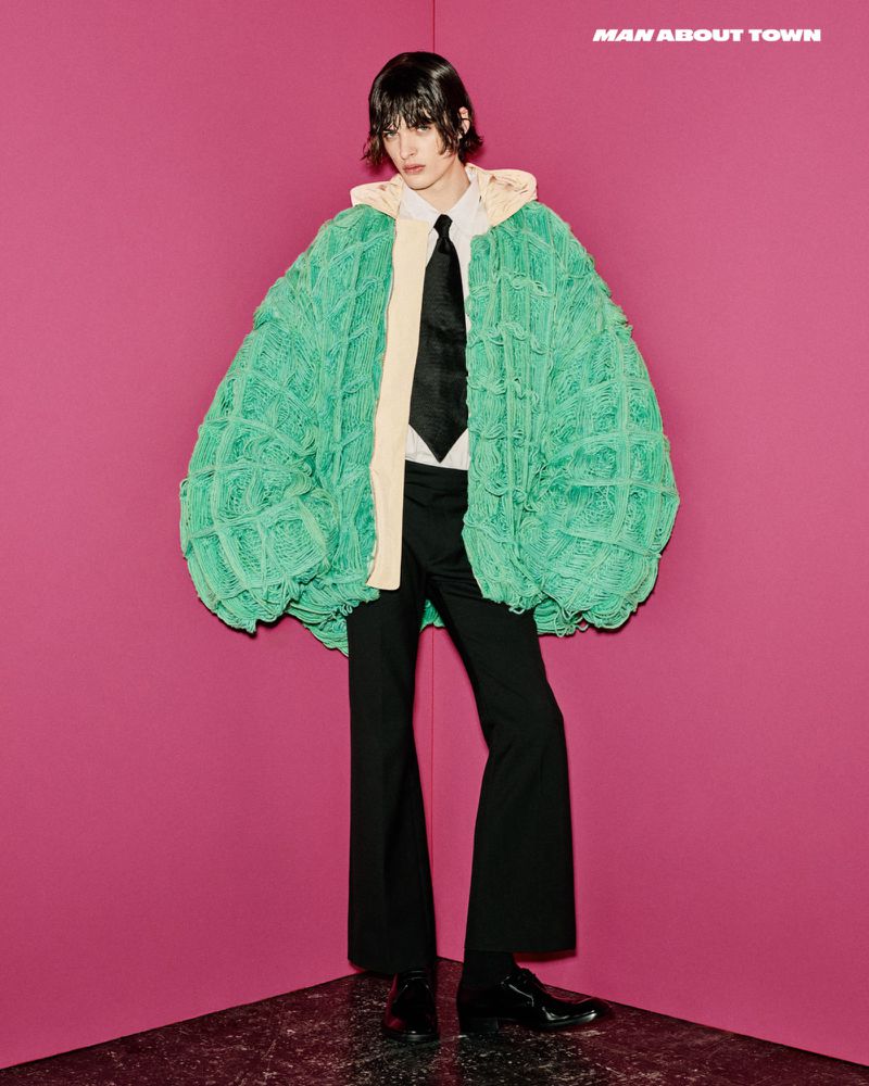 Model wearing an oversized green knit cardigan, white shirt, black tie, and dark trousers against a pink wall.