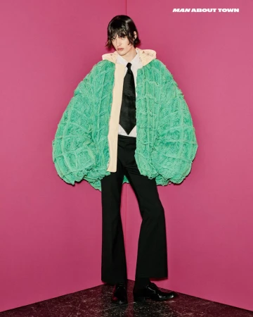 Model wearing an oversized green knit cardigan, white shirt, black tie, and dark trousers against a pink wall.