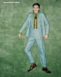 A young male model lying down on a green carpet wearing a light blue suit, green shirt, patterned tie, and dark dress shoes.