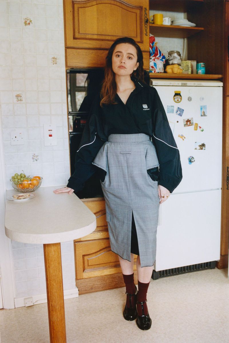 Adult female in a black jacket and grey plaid skirt poses in an indoor kitchen with white appliances and a small table with fruit.
