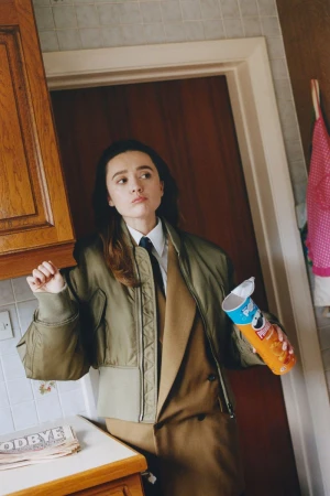 Young adult female poses indoors wearing a khaki bomber jacket, tan suit, and tie; holding a Pringles can in a kitchen setting.