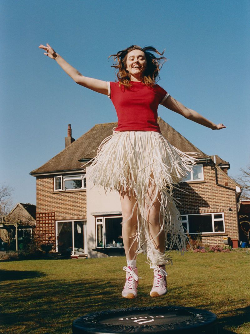 A cheerful woman in a red shirt and a whimsical hula skirt joyfully jumps on a trampoline against a house and clear blue sky.