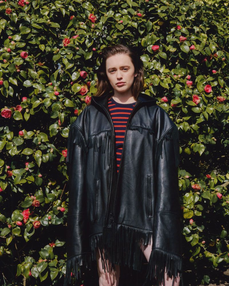 A young woman with short hair stands outside, wearing a black fringed jacket in front of a green leafy bush with red flowers.