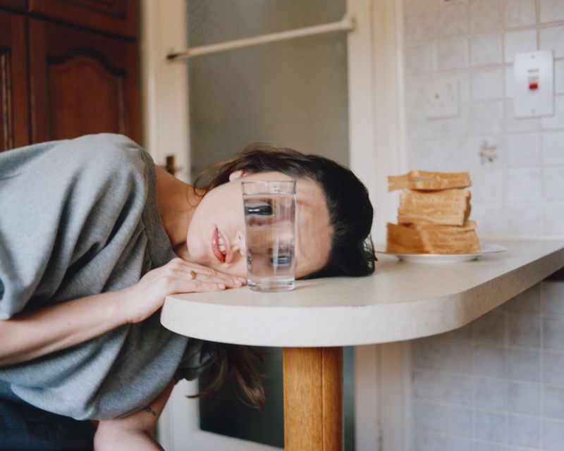 Woman rests head on table with glass of water, stack of toast in kitchen, indoor setting, subdued lighting, medium shot, lifestyle photography.