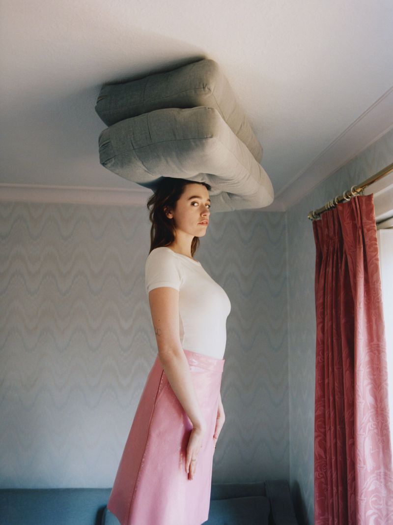 Young woman in white shirt and pink skirt balancing large gray cushions on her head indoors near window with patterned wallpaper.
