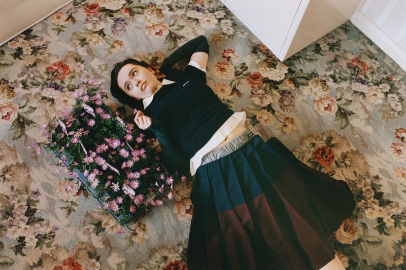 Overhead shot of a woman in a sweater and skirt lying on a floral patterned rug next to a flower basket with pink flowers.
