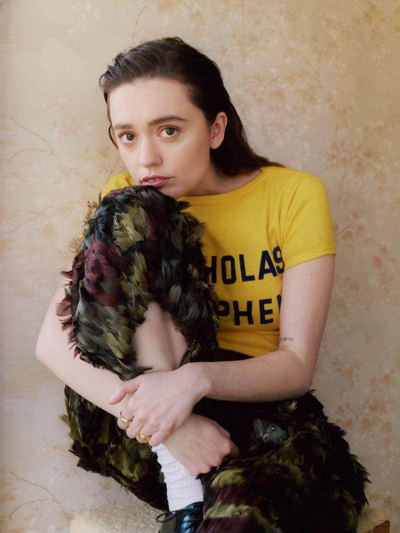 Young woman with dark hair and yellow t-shirt hugs knees covered in feathered fabric in an eye-level indoor studio portrait.